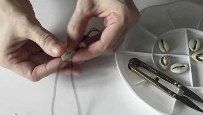 Attach cowrie shells: Thread a cowrie shell onto both cords, creating a cross inside the shell to secure it. Tie a knot at the base of each shell to prevent it from sliding.